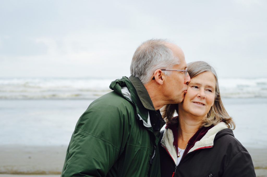 Older man kissing his wife on the cheek on the beach.